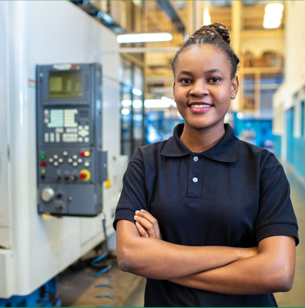 A female technician poses in a factory setting with tecnology behind her.
